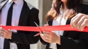Woman in black blazer and white button down shirt cutting a red ribbon with scissors